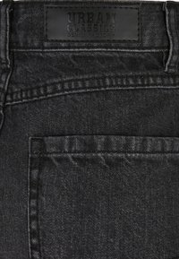 Melni džinsi ir ar ādas etiķeti ar uzrakstu "URBAN CLASSICS DENIM FACTORY", aizmugurē ir kabata un tekstūrētas auduma detaļas.