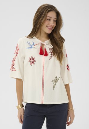 Jauna moteris dėvi baltą tekstūruotą palaidinę su spalvinga paukščių ir gėlių siuvinėjimu bei raudonomis kutomis, derinamą su tamsiomis kelnėmis ir auksinėmis apyrankėmis.