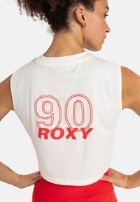 Cropped white sleeveless top featuring large red "90 Roxy" graphic on the back. Soft fabric with a relaxed fit design.