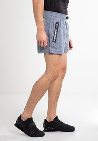 Šedé sportovní kraťasy s texturovaným povrchem, elastickým pasem a bočními kapsami. Černý brandingový štítek na levé straně.