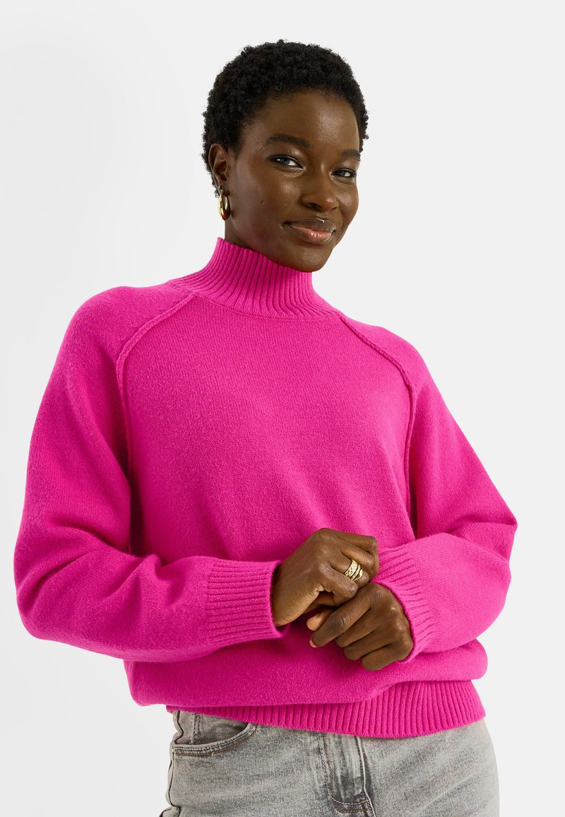 Femme aux cheveux courts et bouclés portant des boucles d'oreilles créoles en or et un pull à col roulé rose vif, posant les mains jointes.