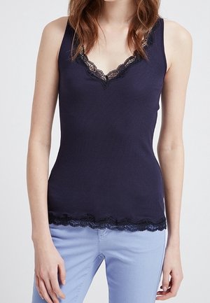 Kobieta ubrana w granatowy top bez rękawów z koronkową lamówką przy dekolcie i na dole, w połączeniu z jasnoniebieskimi spodniami.