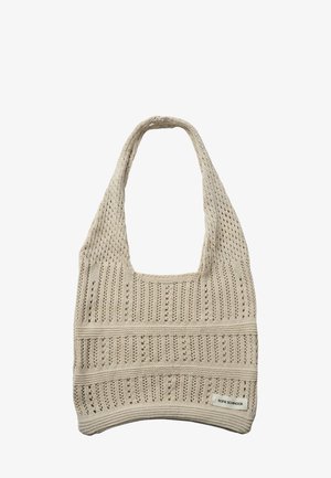 Beżowa dziergana torba typu tote z wzorem ażurowym, szerokim paskiem i małą prostokątną metką z nazwą marki w prawym dolnym rogu.