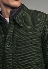 Grünes kariertes Jackett mit spitzem Kragen, einer Brusttasche und schwarzen Knöpfen, über einem grauen Reißverschluss-Hemd getragen. Textilelemente sind sichtbar.