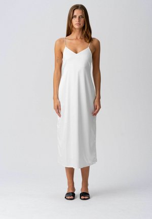 Femme aux longs cheveux bruns portant une robe midi blanche sans manches et des sandales noires à enfiler, debout devant un fond gris uni.