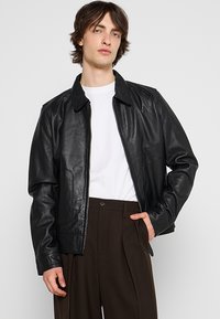 Veste en cuir noir avec une texture lisse, dotée d'un col, d'une fermeture éclair et de poches latérales, portée sur une chemise blanche.