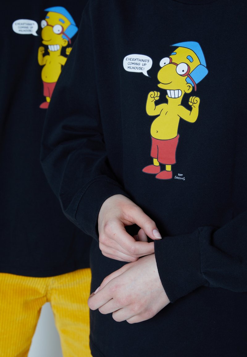 T-shirt noir à manches longues avec un personnage de dessin animé portant un chapeau bleu et un short rouge. La bulle de texte dit : "Tout va pour le mieux, Milhouse."