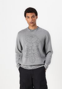 Gray sweater with a floral pattern, featuring a textured knit and a contrasting patterned collar. Worn with black cargo pants.