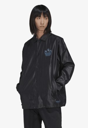 Veste noire, brillante, avec une fermeture à bouton à l'avant, des poches latérales et un logo brodé bleu sur la poitrine gauche. Matériel lisse et léger.