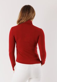 Maglietta rossa a maniche lunghe con scollo a dolcevita, dal design aderente e texture a coste, caratterizzata da un alto colletto e tessuto morbido. Indossata con pantaloni bianchi.