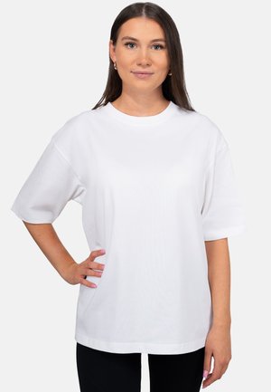 Jonge vrouw met lang bruin haar die een eenvoudige witte oversized T-shirt draagt, staand met één hand op haar heup.