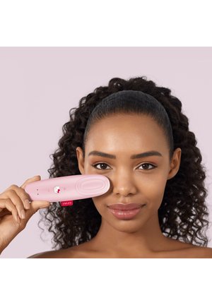 Woman with curly hair holding a pink handheld device with Hello Kitty logo near her cheek, against a plain light purple background.