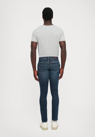 Uomo in piedi con le spalle rivolte, indossa una maglietta grigia chiara, jeans skinny blu scuro e sneakers bianche, davanti a uno sfondo bianco semplice.