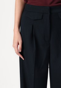 Navy high-waisted trousers with prominent pleats, a smooth texture, and angled pockets, paired with a burgundy top.