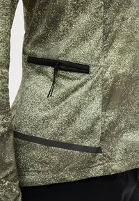 Gros plan d'une personne portant une veste verte mouchetée avec une petite poche zippée et une bande réfléchissante horizontale près de l'ourlet.