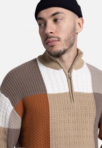 Suéter de punto con diseño de patchwork en colores marrón, beige, blanco y óxido, con un cuello alto y cierre de cremallera. Patrones texturizados visibles.