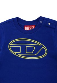 T-shirt in cotone blu con scollo rotondo, caratterizzato da un grande logo ovale giallo. Dettaglio con chiusura a pressione sulla spalla. Etichetta del brand rossa visibile.