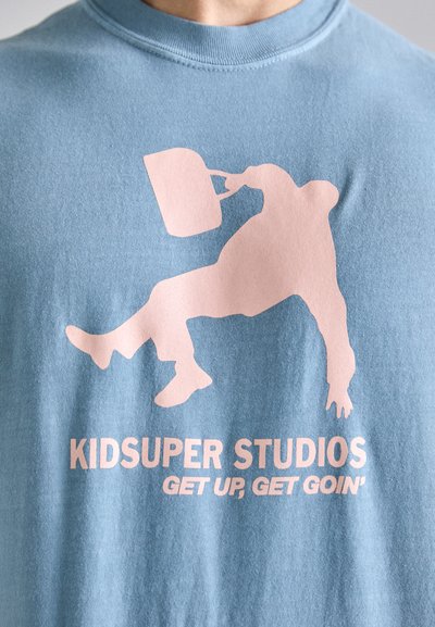 Hele sinise puuvillase T-särk, millel on roosa graafika inimesest liikumises, kes hoiab konteinerit, allpool on tekst "KID SUPER STUDIOS".