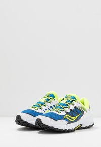Blaue und weiße Sportschuhe mit schwarzen Sohlen, ausgestattet mit einem Mesh-Obermaterial, gelben Akzenten und kontrastierenden Schnürsenkeln, die für Griffigkeit konzipiert sind.