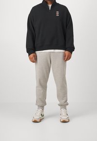 Felpa nera con zip e loghi ricamati, pantaloni felpati grigi e sneakers bianche con dettagli verdi e beige su uno sfondo neutro.