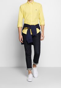 Żółta koszula na guziki z długim rękawem i kołnierzykiem. Granatowa bluza z kapturem związana w talii. Ciemnoniebieskie dopasowane jeansy i białe trampki.