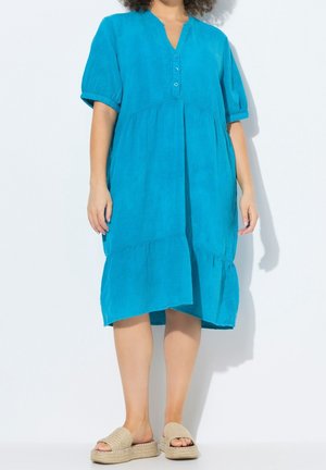 Femme portant une robe turquoise ample longueur genou avec des boutons et des manches bouffantes, associée à des sandales beige à plateforme.