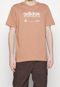 Krátký rukáv, tričko z broskvového bavlněného materiálu s bílým textem a logem "adidas SPORTSWEAR" tištěným na přední straně. Hladký povrch, kulatý výstřih.
