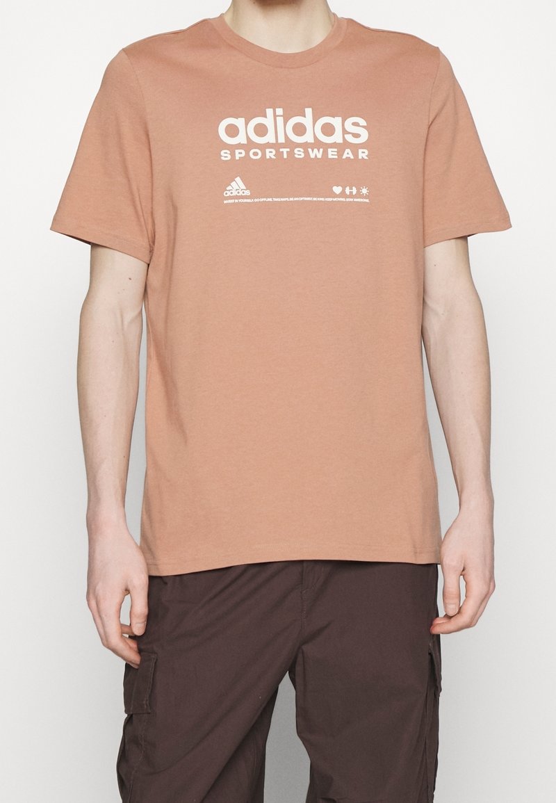 Krátký rukáv, tričko z broskvového bavlněného materiálu s bílým textem a logem "adidas SPORTSWEAR" tištěným na přední straně. Hladký povrch, kulatý výstřih.