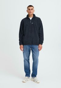 Pullover in pile nero con zip a quarti, superficie texturizzata e colletto alto. Indossato con jeans azzurri e scarpe di colore chiaro.
