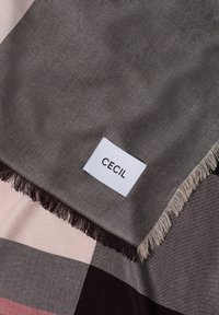 Tissu à franges marron et beige avec une étiquette blanche indiquant "CECIL", placée près du bord du tissu sur une surface à carreaux.
