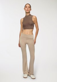 Top corto marrone con colletto alto e design aderente, abbinato a pantaloni beige a campana. L'outfit presenta una texture liscia e una leggera lucentezza.