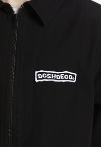 Chaqueta negra con cremallera y tejido texturizado; presenta un logo bordado en blanco "DCSHOECO" en el lado izquierdo del pecho.