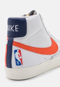 Bílé kožené vysoké Nike tenisky s oranžovým swooshem a modrými akcenty. Obsahují nášivku NBA, texturovanou podrážku a prošívané detaily.