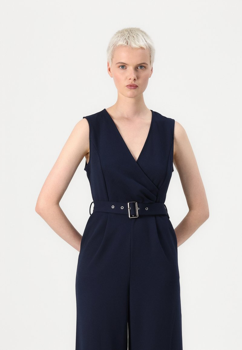 Tuta navy con scollo a V profondo e design senza maniche. Presenta una vita aderente con una cintura larga e una fibbia quadrata in metallo. Tessuto dalla consistenza liscia.