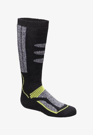 Calcetines de esquí negros con rayas grises y amarillas, con un diseño acolchado. Un niño con una chaqueta azul está sentado en un banco de madera con botas de esquí.