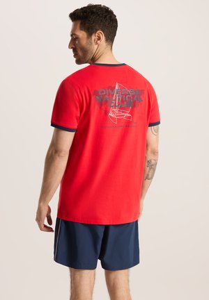 Czerwona bawełniana koszulka z granatowym wykończeniem, z nadrukiem łódki żaglowej oraz napisem "Diverse Nautical Club" na plecach. Noszona z ciemnymi szortami.