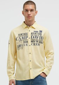 Helles gelbes Langarmshirt mit navyfarbenem Grafiktext und Aufnähern, Knopfverschluss und kontrastierenden Nähten an den Ärmeln.