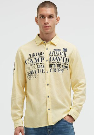 Light yellow long-sleeve shirt featuring navy graphic text and patches, button closure, and contrasting stitching detail on sleeves.