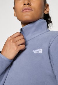 Persona, kas aizver gaiši zilu polārjaka rāvējslēdzēju ar "The North Face" logotipu uz krūtīm uz viendabīga fona.