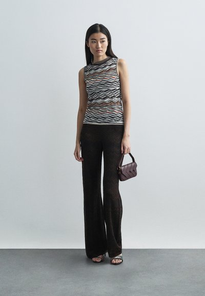 Kvinna i ärmlös mönstrad topp, genomskinliga svarta zigzagbyxor, sandaler, som håller i en liten mörk handväska, stående mot en enkel grå bakgrund.