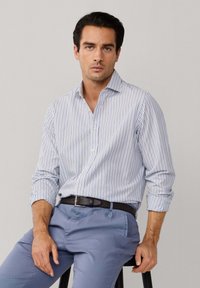 Muškarac s tamnom kosom, obučen u svijetloplavu i bijelu prugastu košulju s gumbima i plave hlače, sjedi na crnoj stolici na jednostavnoj pozadini.