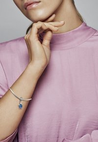 Bracciale in argento con pendente in gemma blu, caratterizzato da una texture liscia e dettagli intricati, indossato su una mano su sfondo rosa pastello.