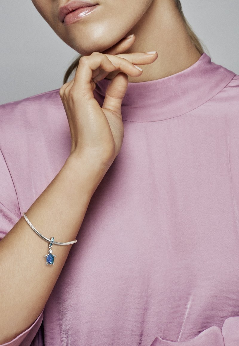 Bracciale in argento con pendente in gemma blu, caratterizzato da una texture liscia e dettagli intricati, indossato su una mano su sfondo rosa pastello.