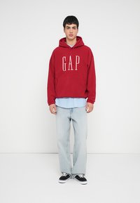 Felpa con cappuccio rossa con logo "GAP", abbinata a una camicia a righe blu sottostante, jeans azzurri e sneaker nere.