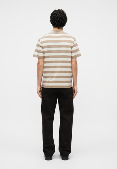 Άνδρας με σγουρά μαλλιά που φοράει μπεζ και λευκό ριγέ κοντομάνικο πουκάμισο και μαύρο παντελόνι, στέκεται με την πλάτη στραμμένη σε μονόχρωμο φόντο.