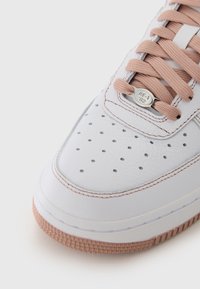 Weiße Ledersneaker mit strukturierter Oberfläche, pinken Schnürsenkeln, Belüftungsperforationen und einer hellpinkfarbigen Gummisohle. Sichtbares metallisches Logodetail.