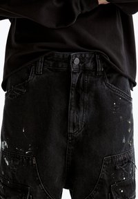 Pantalones de denim negro con detalles desgastados, salpicaduras de pintura visibles y cierre estándar con botón en la cintura. Incluyen bolsillos laterales.