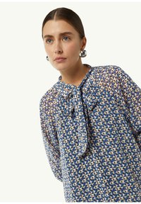 Durchsichtige blaue Bluse mit geometrischen Mustern, einem Bindekragen und lockeren Ärmeln, die eine Mischung aus hellen und dunklen Farbtönen zeigt.