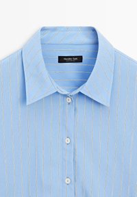 Lyseblå skjorte med tynde hvide lodrette striber, spids krave og hvide knapper foran.