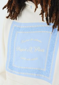 Felpa crema con stampa paisley azzurra e testo ricamato, con un design rettangolare e bordo dettagliato. Tessuto morbido.